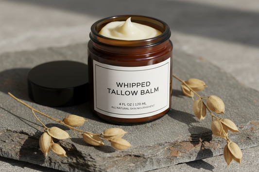 Whipped grass-fed tallow balm in 4oz amber glass jar with white label and black lid, displayed with jojoba plant sprigs on natural stone